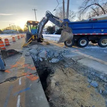 Backhoe loading dirt into a dump truck at a road construction site with orange traffic cones.