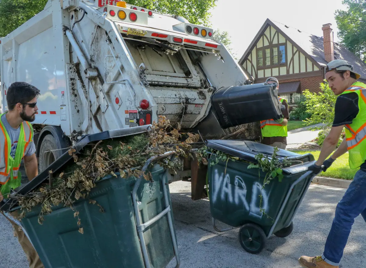 Two sanitation workers in high-visibility vests are emptying yard waste bins into the back of a garbage truck on a
