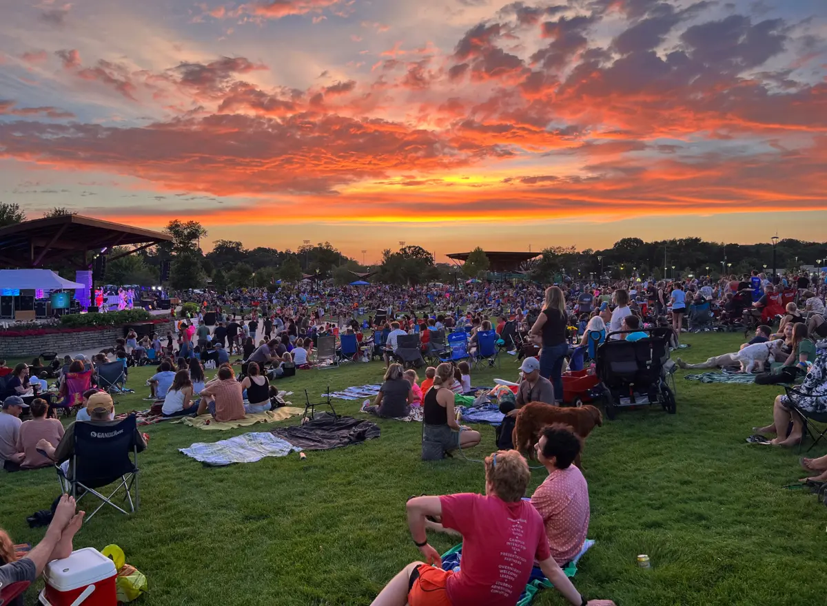 A large crowd gathers on a grassy lawn to watch a performance on an outdoor stage, silhouetted against a vibrant sunset sky.