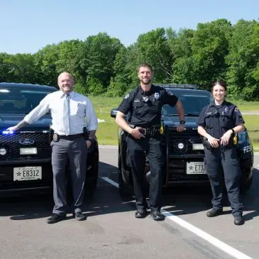 Three police officers stand between two police vehicles. The officer on the left wears a light blue shirt and grey pants.