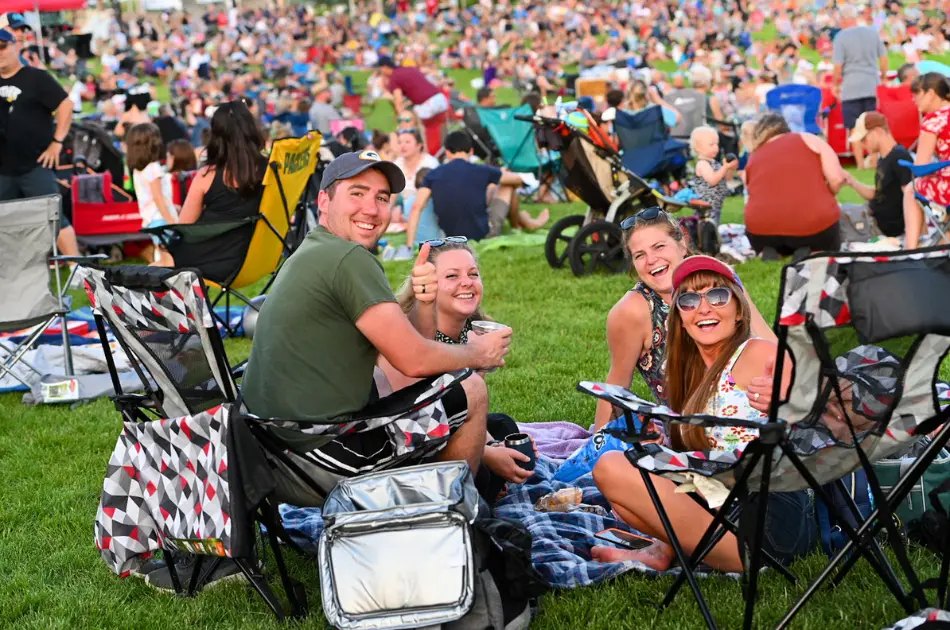 Four smiling people sit on blankets and chairs on a grassy lawn, with a large crowd in the background at an outdoor event.