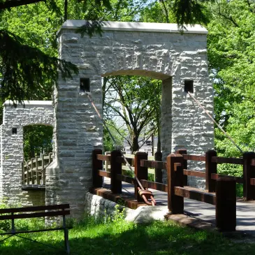 Stone suspension bridge with wooden railings and cables, surrounded by lush green trees and foliage.