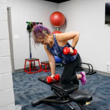 Woman performing a dumbbell row exercise on a weight bench, with fitness equipment in the background.