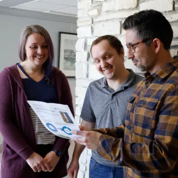 Three colleagues review a document with charts and text, smiling and engaged.