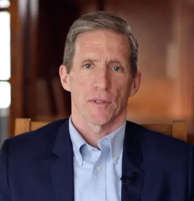 A man in a navy suit and light blue collared shirt looks directly at the camera.