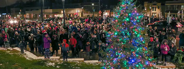  A large crowd gathers at night around a brightly lit Christmas tree in a town square.