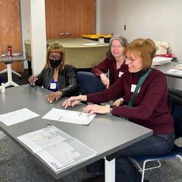 Three women sit at a table reviewing documents, likely poll workers preparing for an election.