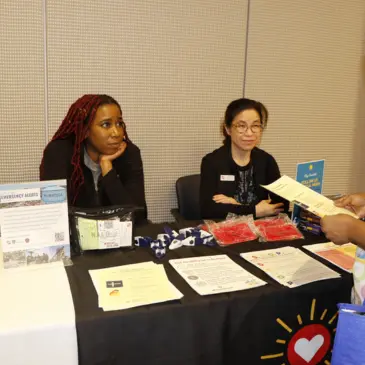 Two women sit behind a table at an event, with informational materials and promotional items displayed.
