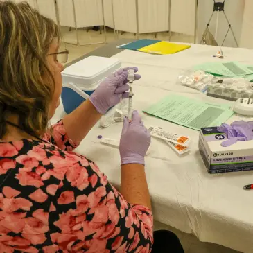 Healthcare worker in purple gloves prepares a syringe with vaccine from a vial at a table with medical supplies.