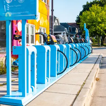 A bright blue bike rack with several bicycles docked. A colorful mural is visible on a building in the background.