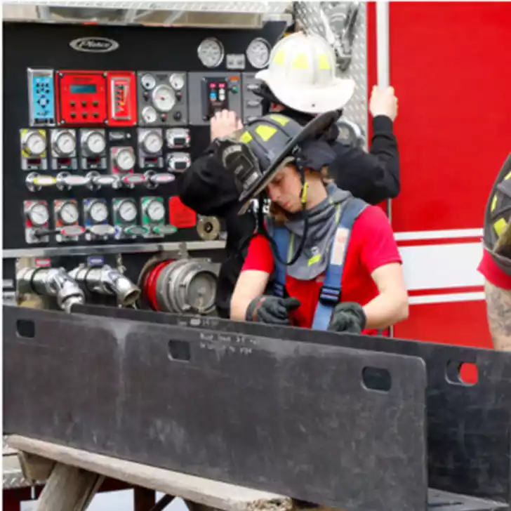 Firefighters in helmets and gear work on the control panel of a fire truck, with gauges and hoses visible.