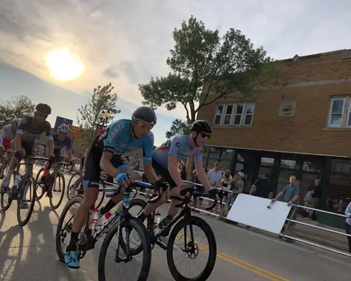 Cyclists in a race, wearing racing jerseys and helmets, with buildings and spectators in the background.