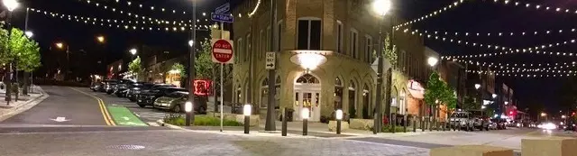  Street scene at night with buildings, parked cars, streetlights, and decorative string lights.