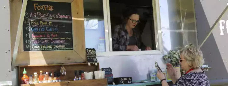 A woman orders from a food truck with a menu board listing items like "Hot Fried Chicken Sandwich" and "Grilled Cheese."