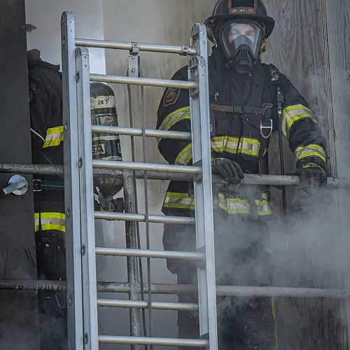 Firefighter in full gear, including mask and helmet, stands near a ladder amidst smoke.