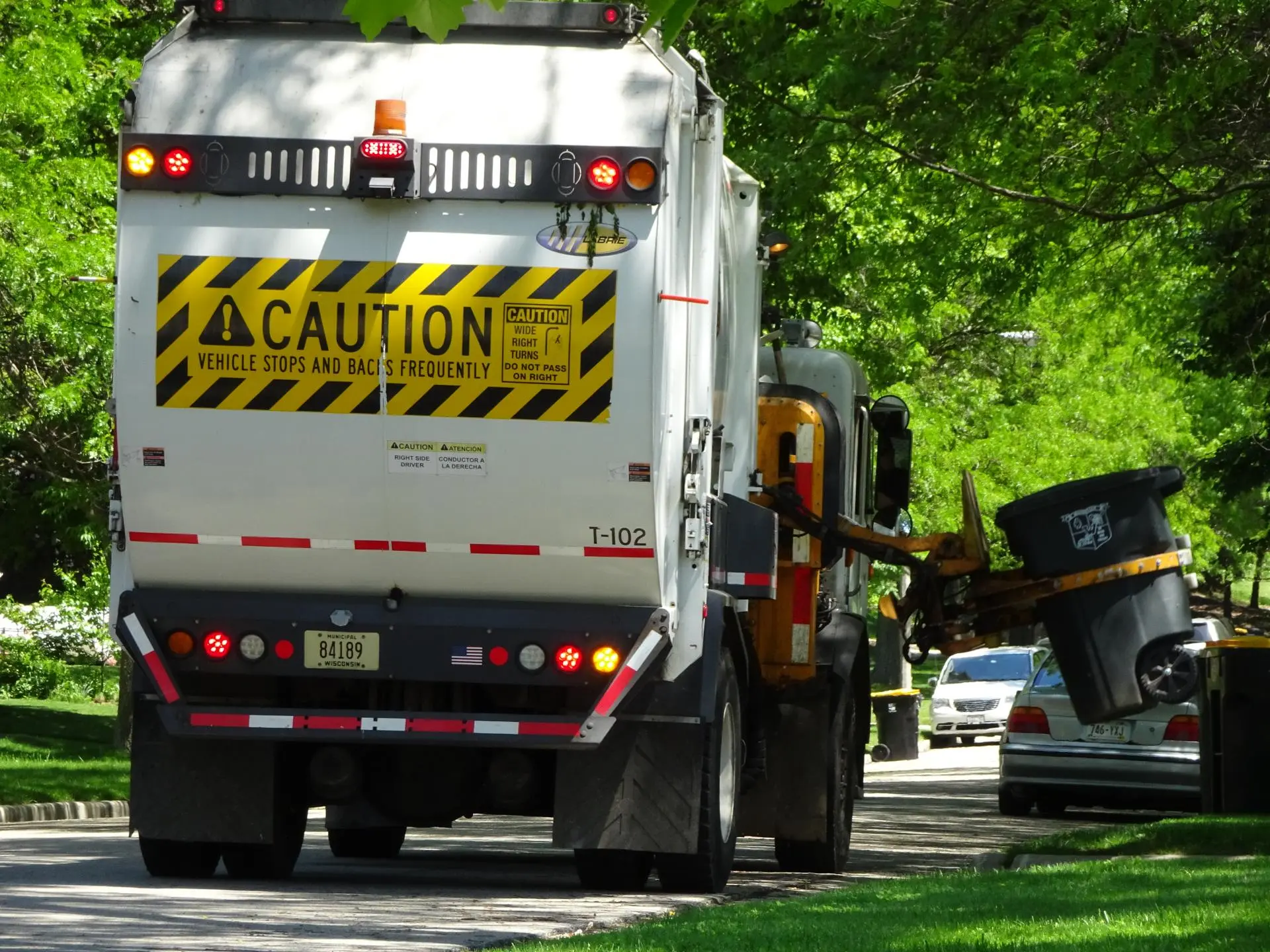 Rear view of a garbage truck with a "CAUTION VEHICLE STOPS AND BACKS FREQUENTLY" sign and a trash bin being lifted.
