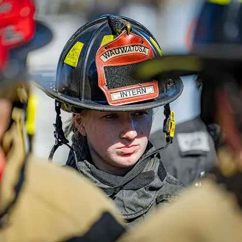 A female firefighter intern wearing a Wauwatosa helmet looks intently forward, surrounded by other firefighters.