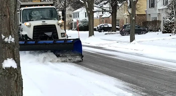 A white snow plow truck with a blue plow attachment clears snow from a residential street during a snowstorm.