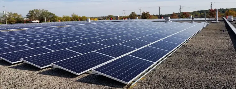 Rows of solar panels installed on a flat, gravel-covered rooftop under a clear sky.