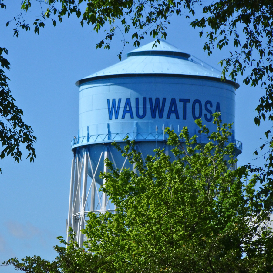 A bright blue water tower with "WAUWATOSA" written in large blue letters against a clear blue sky, framed by green leaves.