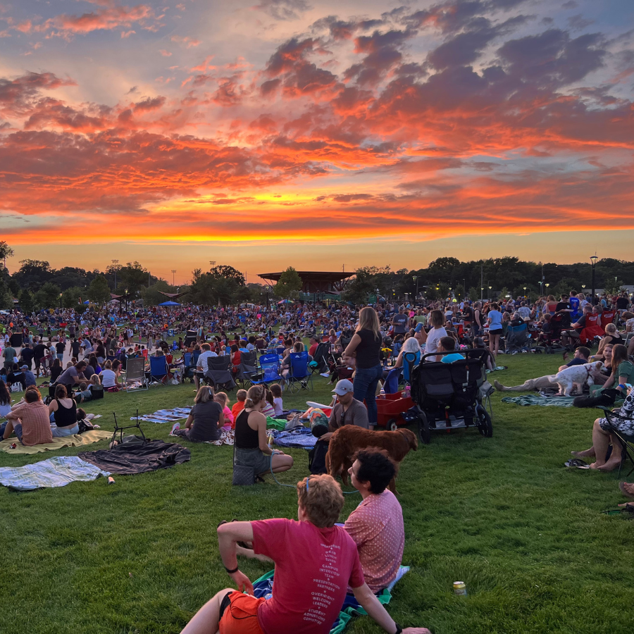 A large crowd gathers on a grassy field watching a vibrant sunset with fiery orange and pink clouds.