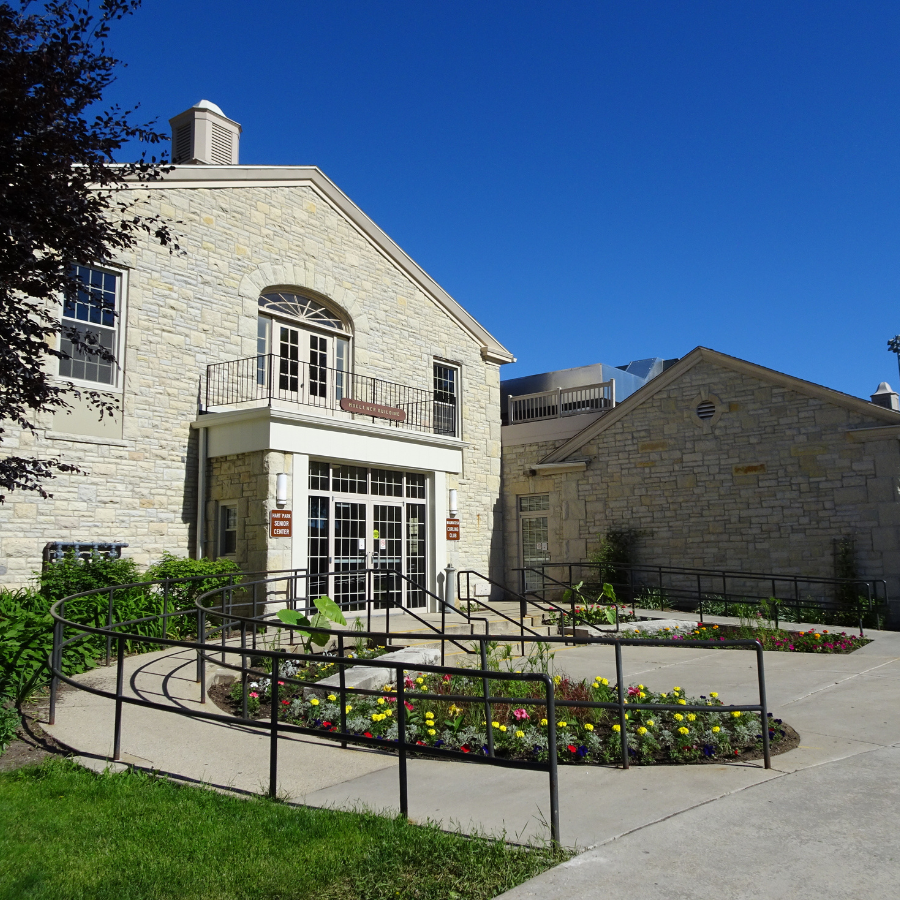 Stone building with a prominent entrance featuring double doors, a balcony above, and a curved walkway with flower beds.