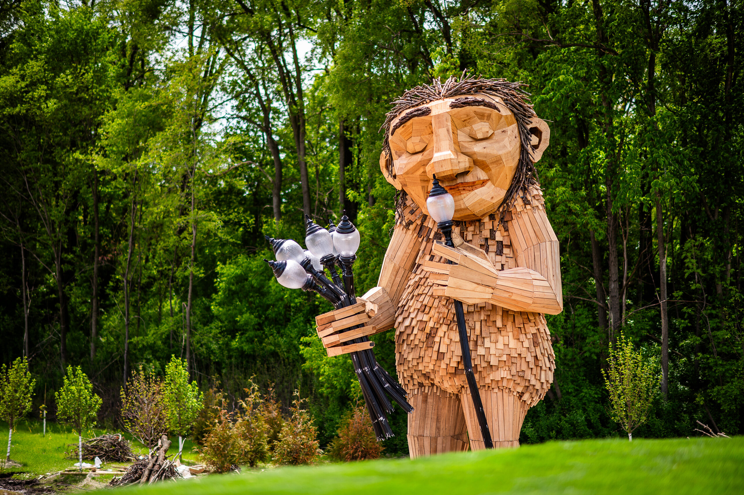 Large wooden sculpture of a troll holding a cluster of lamps and a walking stick in a park.