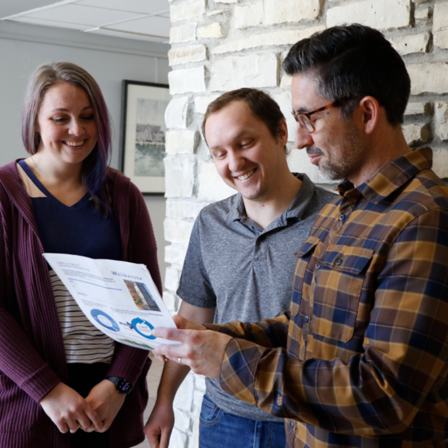 Three colleagues, two men and one woman, smiling and looking at a document with charts and graphs.