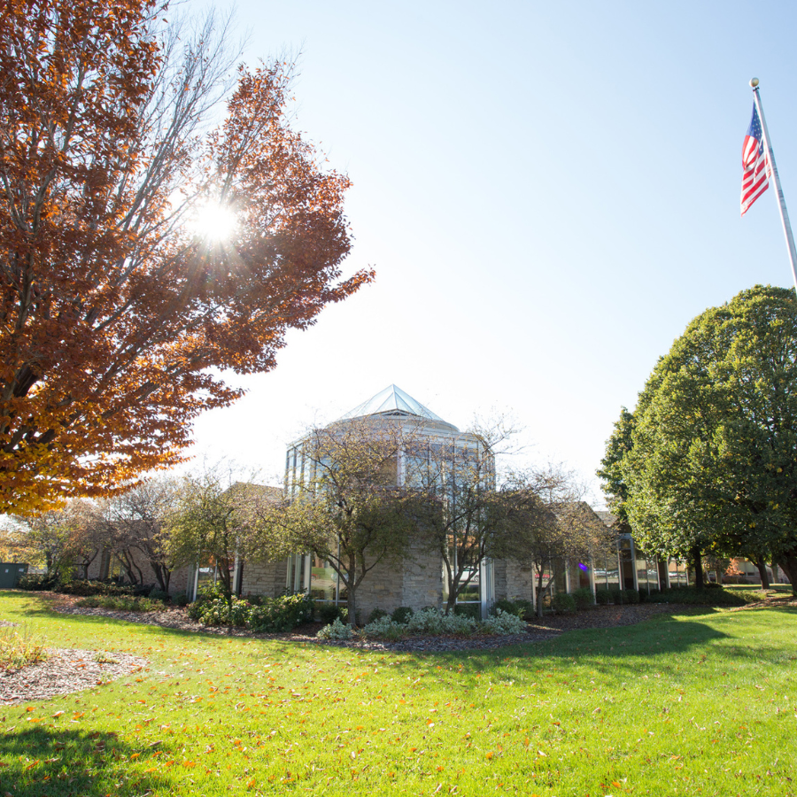 Modern stone building with a glass dome roof, surrounded by trees in autumn. An American flag flies nearby.