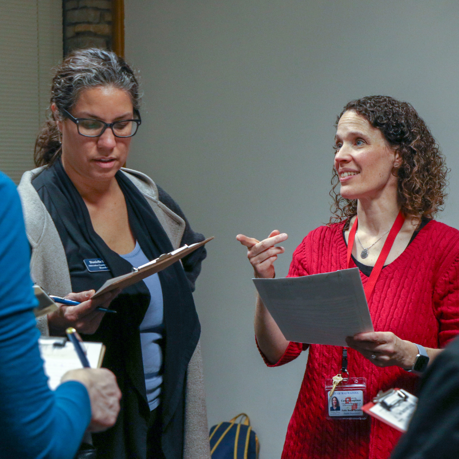 Two women in a meeting, one holding a clipboard and the other gesturing while holding papers.