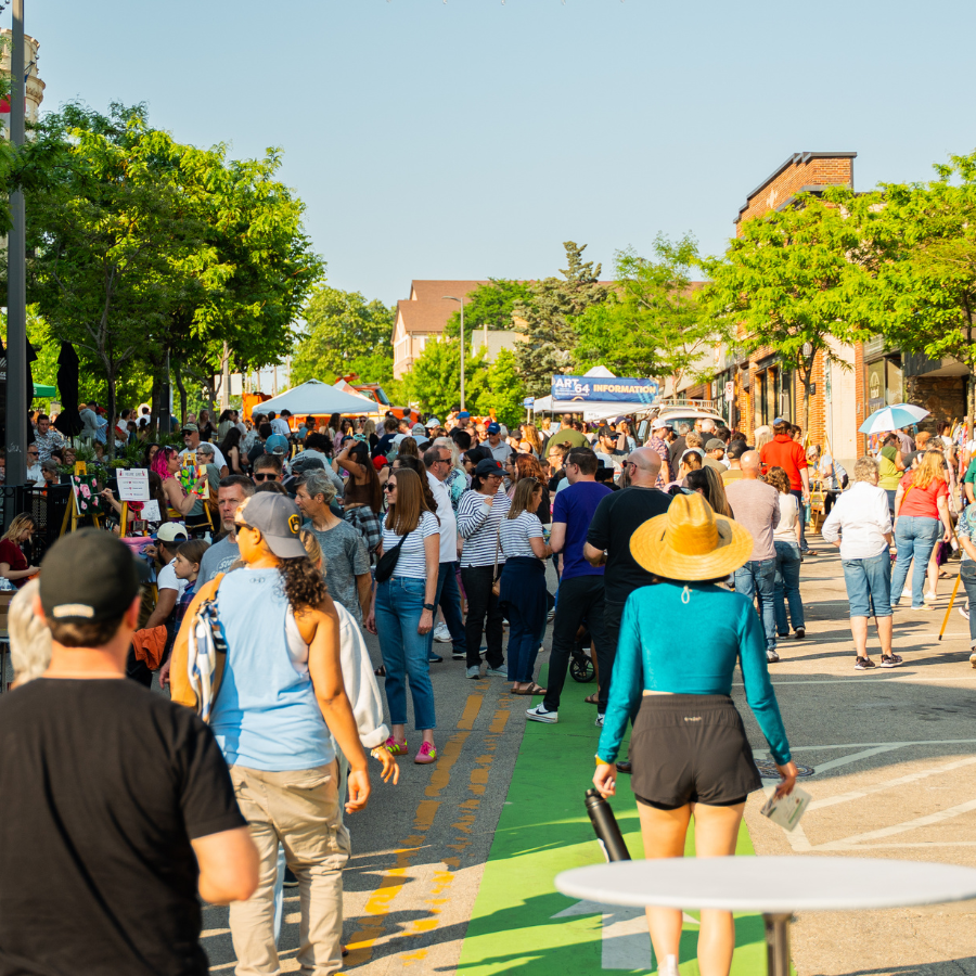 A bustling street fair with many people walking, browsing stalls, and enjoying the sunny weather.